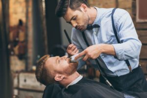 male-barber-trimming-customers-beard-in-barber-sho-FCQHKT7